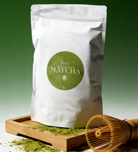 White package of matcha (500g) with a green label on a wooden tray with matcha powder and a whisk.