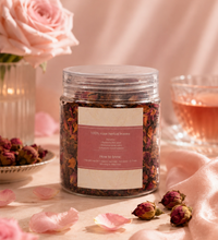 Jar of Rosie Tea with dried rose petals, pink roses, and a glass cup of tea on a soft pink background.