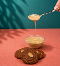 Creamy tahini sauce drizzling into a glass bowl with almond-topped cookies on a pink surface.