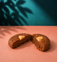 Broken almond-topped chewy tahini cookie on a pink surface with a teal background.