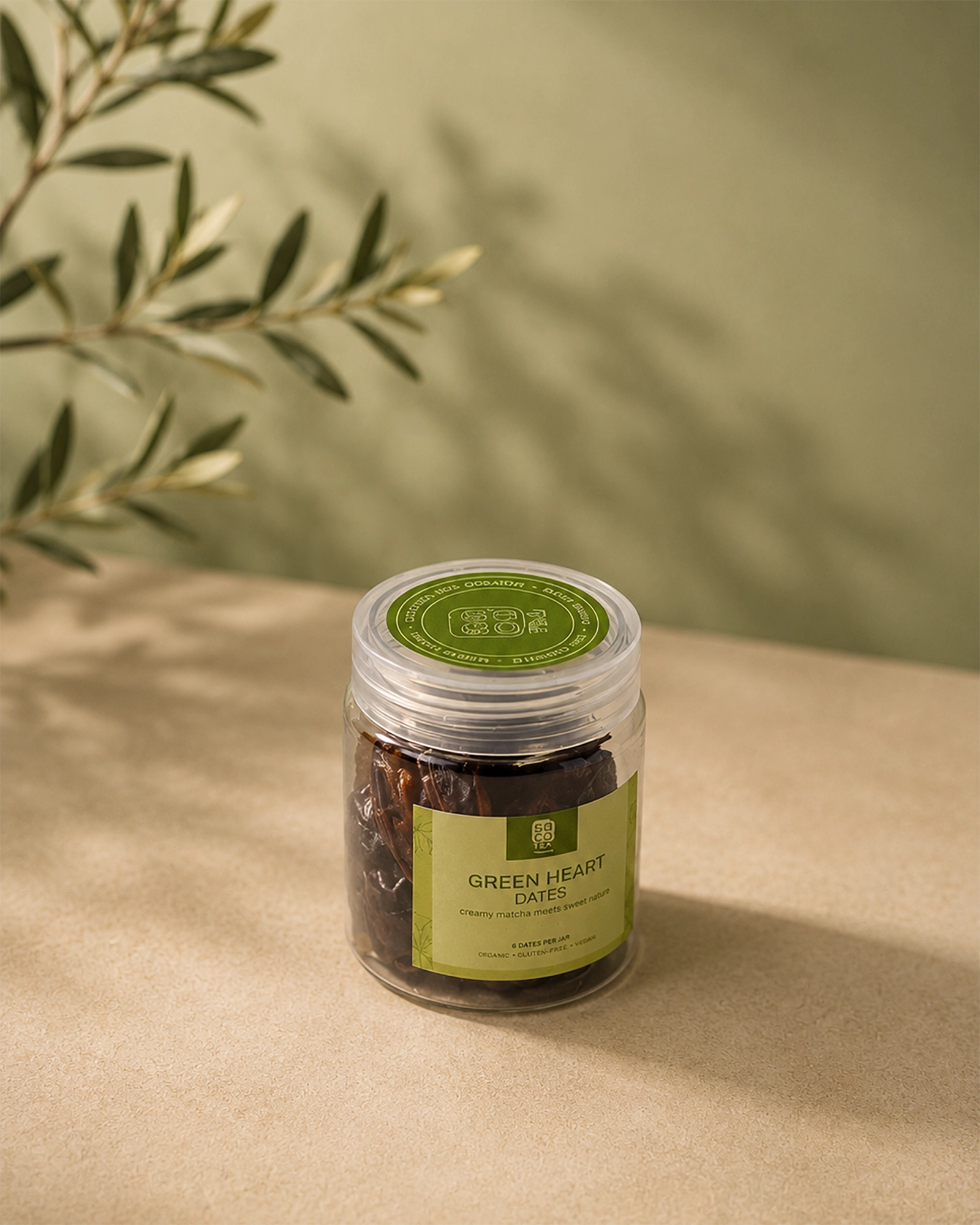 Saco Tea Green Heart Dates jar on a beige surface with soft olive branch shadows.