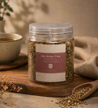 Jar of 'De-Bloa-Tea' with a label on a wooden surface next to a cup and spoon.