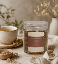 Jar of mixed herbal tea with a label, a ceramic cup, and dried herbs on a neutral background.