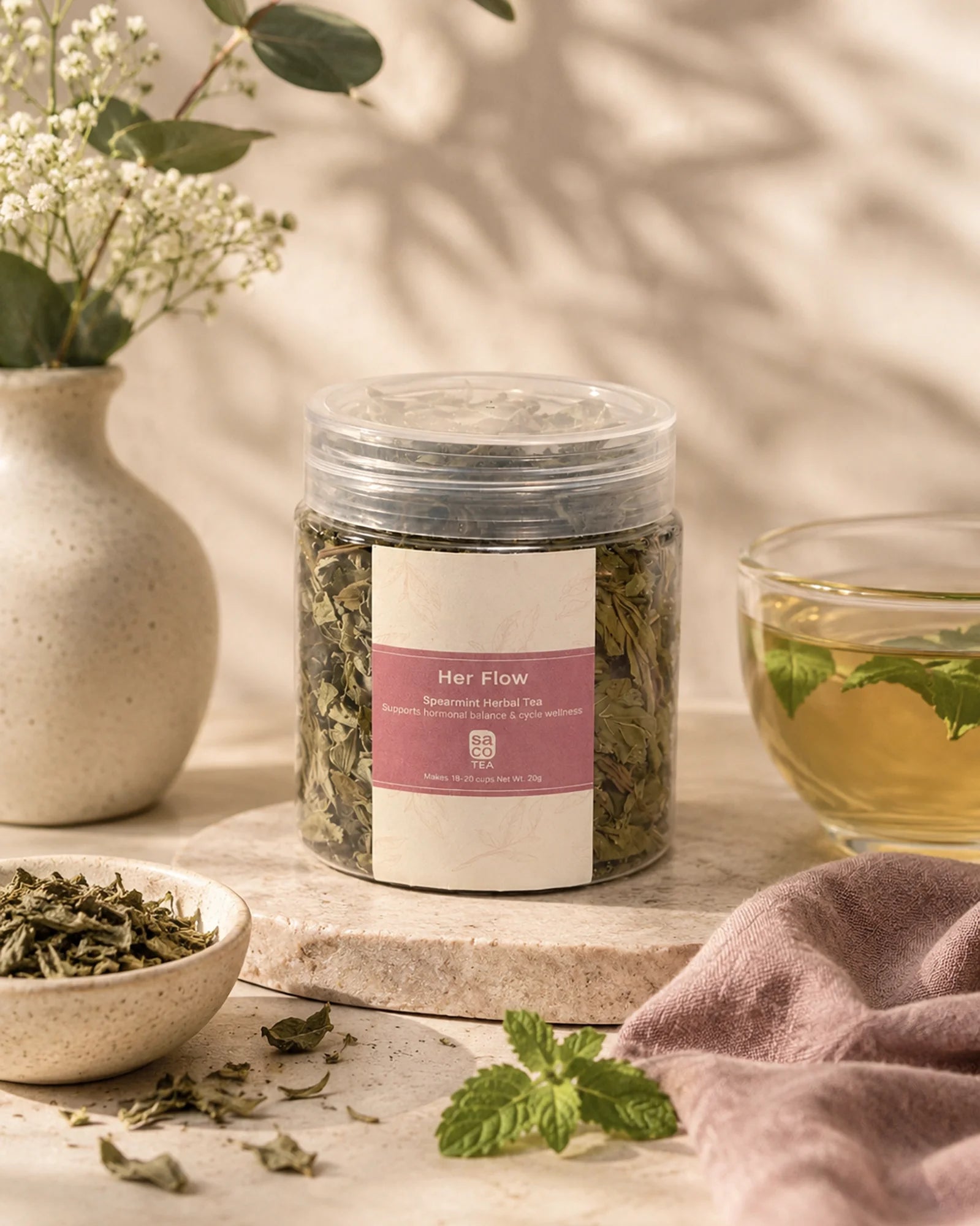 Saco Tea Her Flow herbal tea jar with brewed tea, loose herbs, mint, and soft floral styling on a neutral surface.