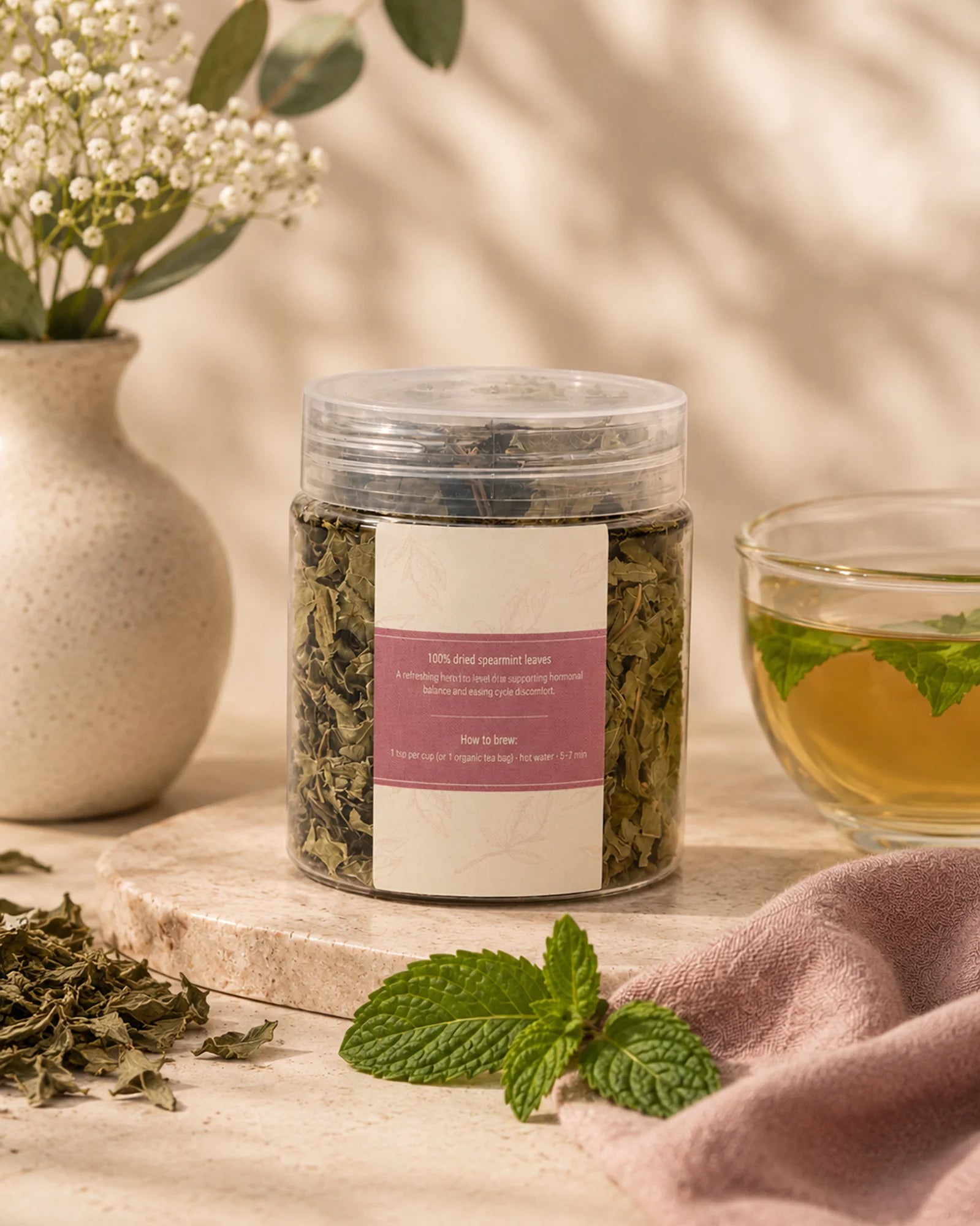 Her Flow herbal tea jar beside a cup of tea, dried herbs, mint leaves, flowers, and a pink cloth on a beige surface.
