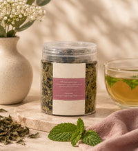 Her Flow herbal tea jar beside a cup of tea, dried herbs, mint leaves, flowers, and a pink cloth on a beige surface.