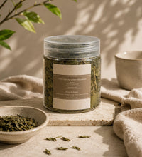 Jar of green sencha tea leaves on a stone tray with loose leaves and a small bowl in soft natural light.