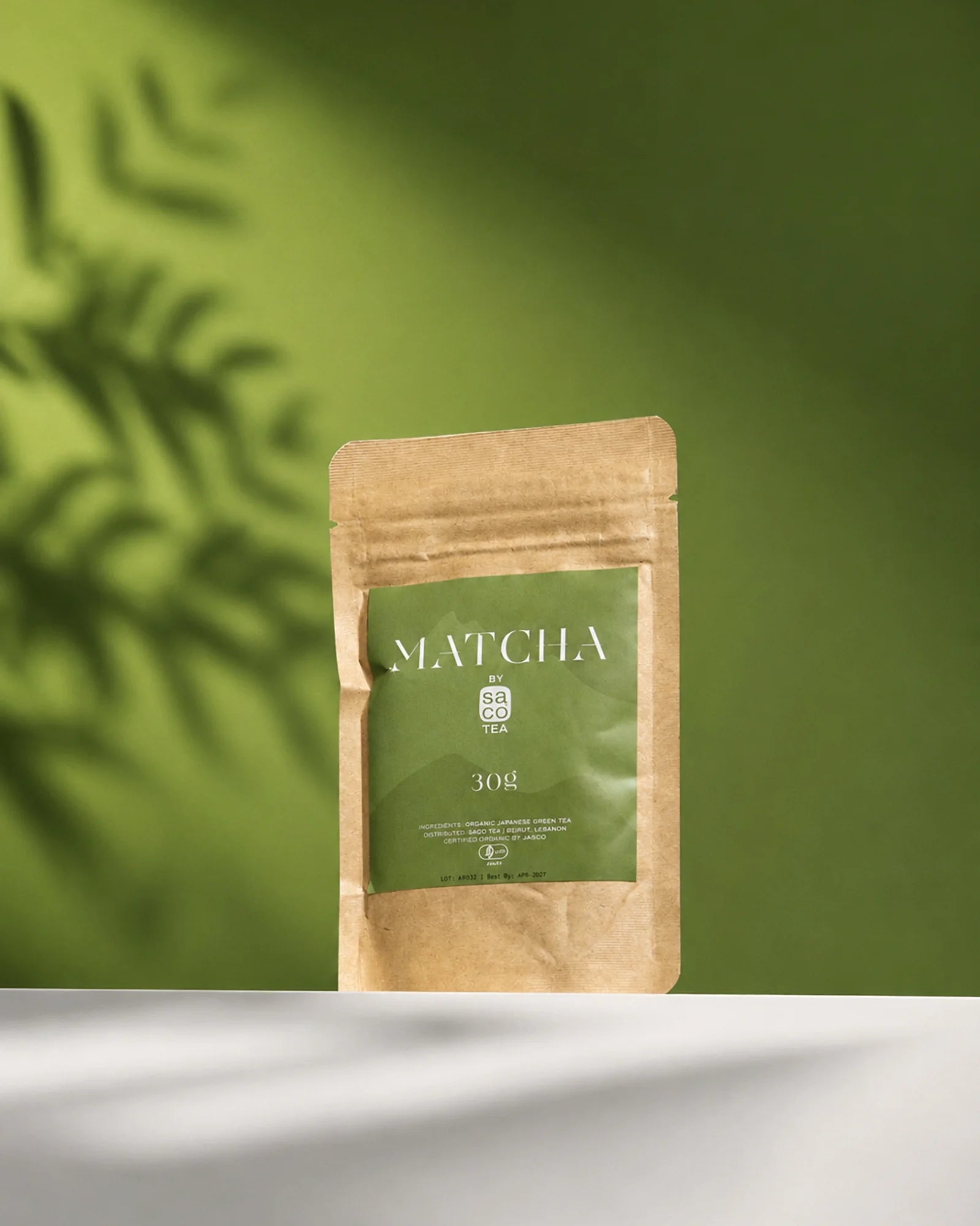 Saco Tea Japanese matcha powder pouch on a green background with soft leaf shadows.