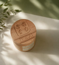 Saco Tea Japanese matcha wooden container on a light surface with soft greenery shadows.