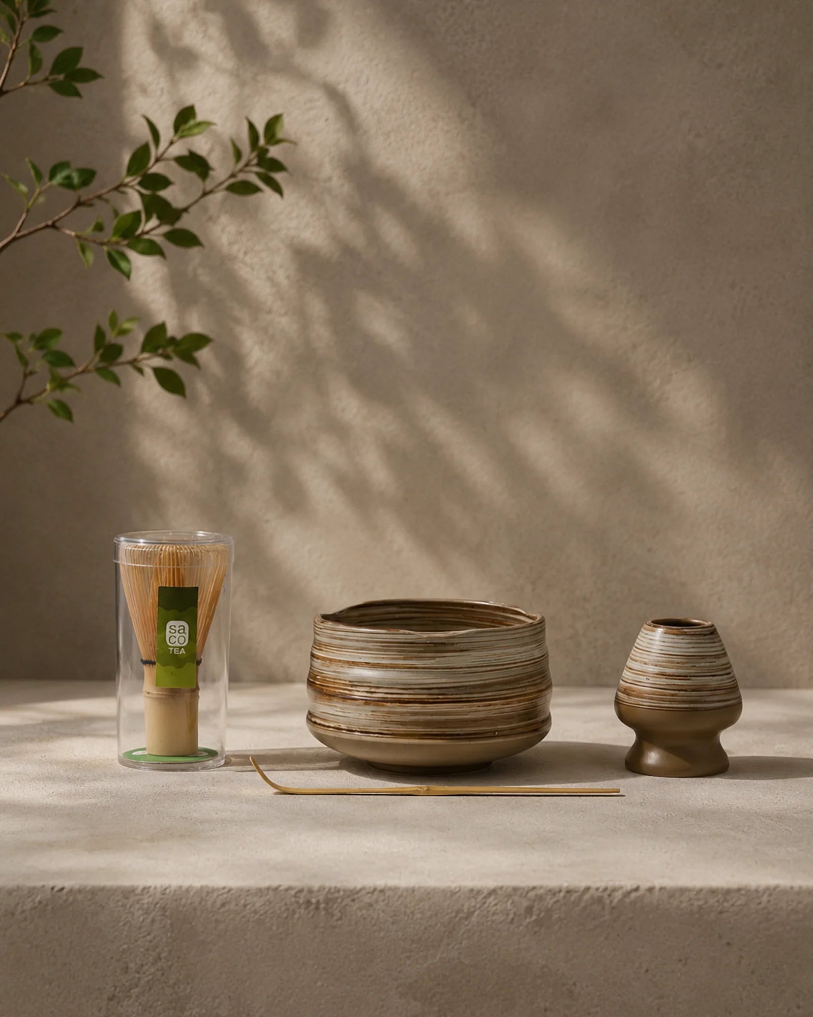 Earthy matcha set with striped ceramic bowl, bamboo whisk in case, ceramic whisk holder, and bamboo scoop on a stone surface.