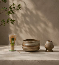 Earthy matcha set with striped ceramic bowl, bamboo whisk in case, ceramic whisk holder, and bamboo scoop on a stone surface.