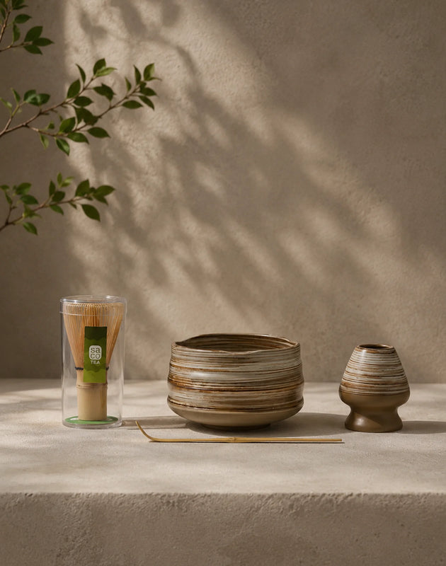Earthy matcha set with striped ceramic bowl, bamboo whisk, ceramic whisk holder, and bamboo scoop on a stone surface.