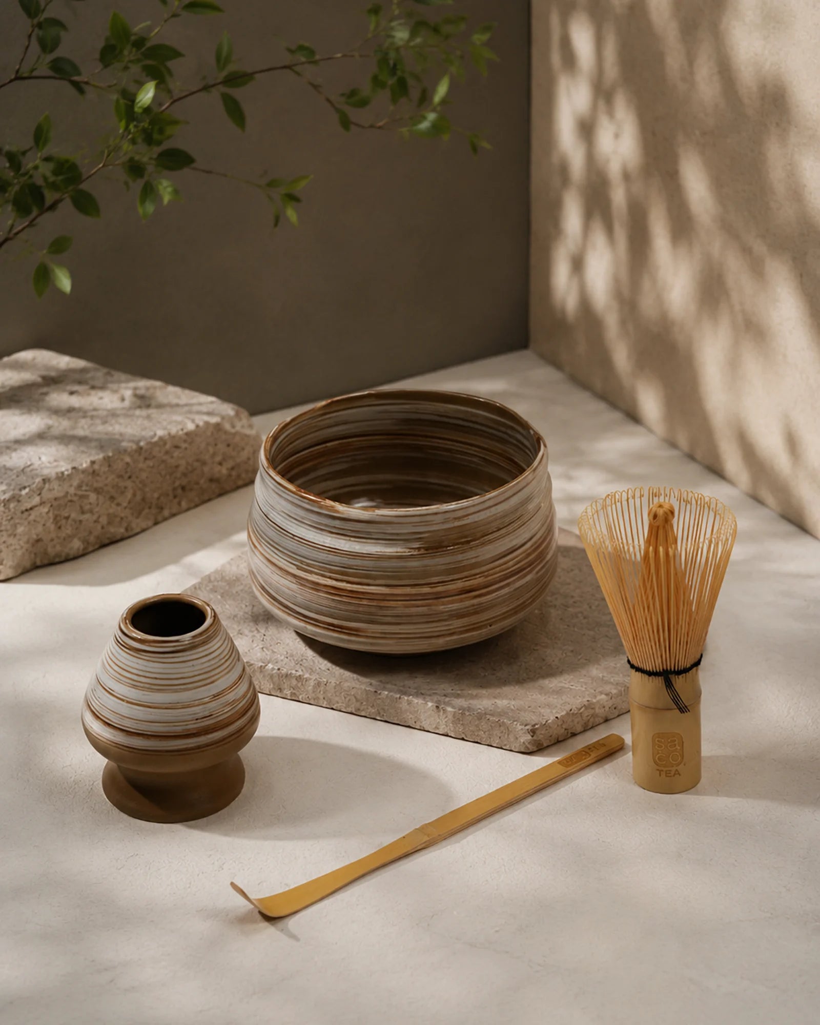 Earthy matcha set with striped ceramic bowl, bamboo whisk, ceramic whisk holder, and bamboo scoop on a stone surface.