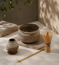Earthy matcha set with striped ceramic bowl, bamboo whisk, ceramic whisk holder, and bamboo scoop on a stone surface.