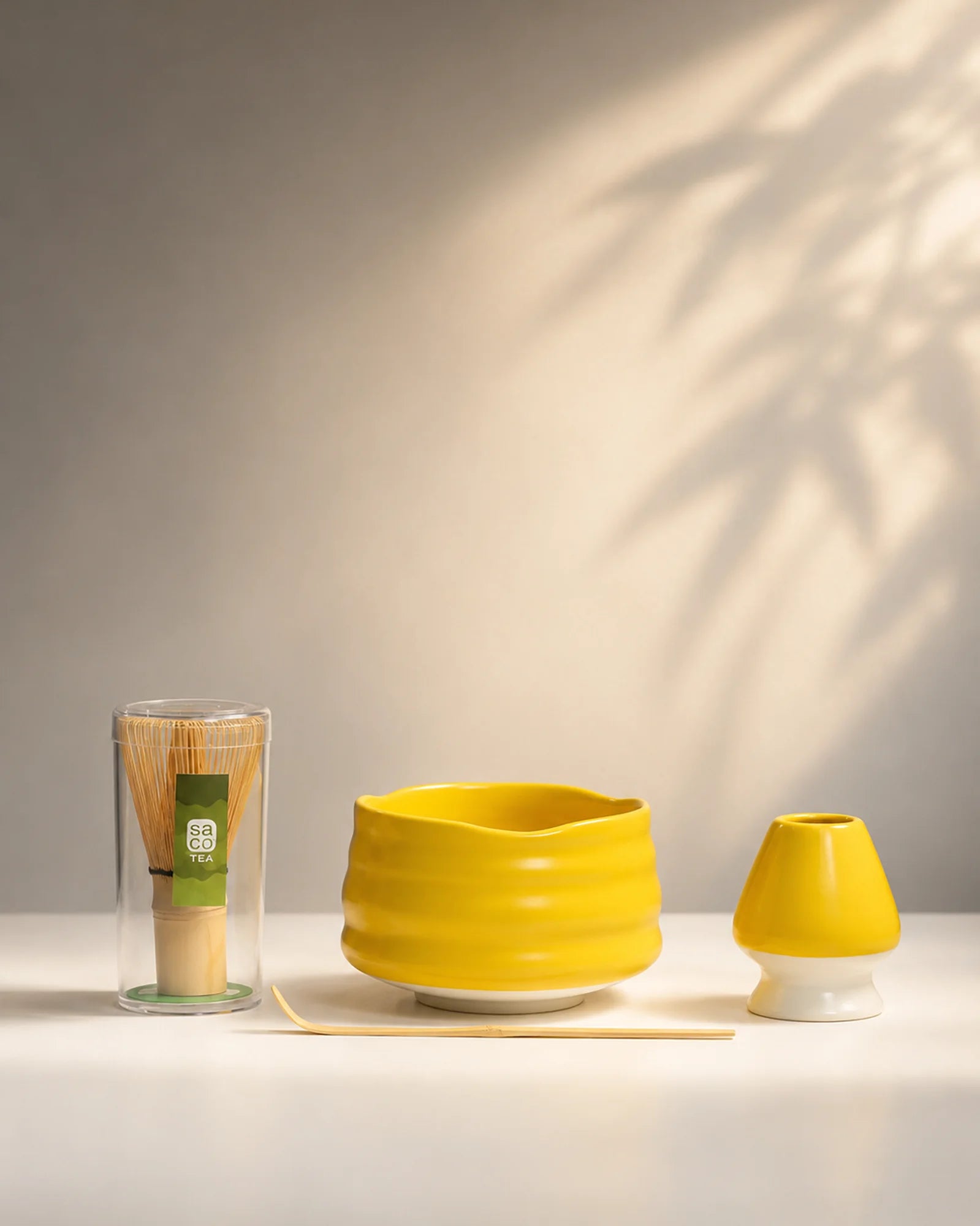 Saco Tea yellow matcha set with ceramic bowl, bamboo whisk in case, whisk holder, and bamboo scoop on a white surface.