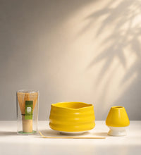 Saco Tea yellow matcha set with ceramic bowl, bamboo whisk in case, whisk holder, and bamboo scoop on a white surface.