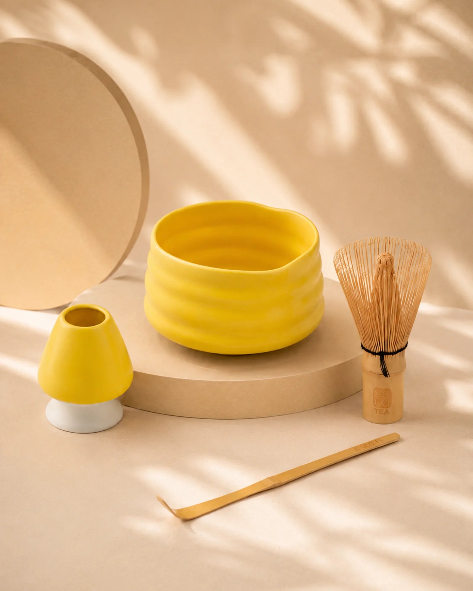 Yellow matcha bowl with bamboo whisk, scoop, and ceramic whisk holder displayed under soft natural shadows.