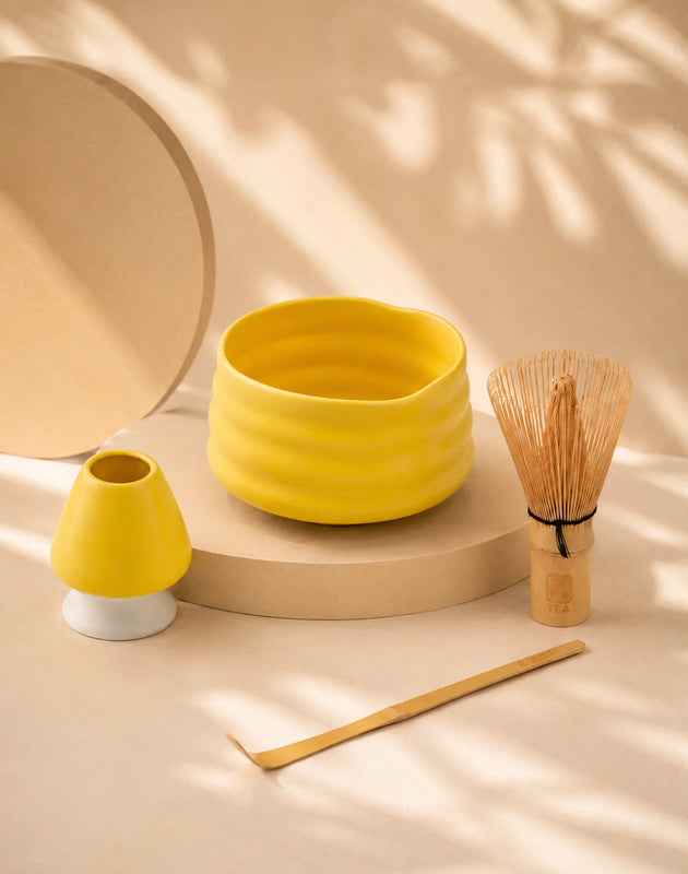 Yellow matcha bowl with bamboo whisk, scoop, and ceramic whisk holder displayed under soft natural shadows.
